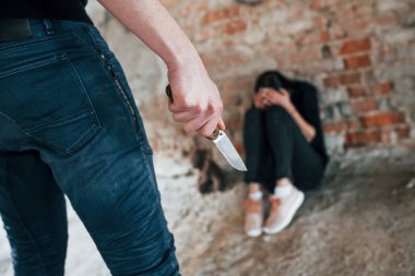 Violent man standing with knife in hand and threatens girl that sits on the floor in abandoned building.