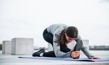 Genç sporcu kız gündüz vakti göl kenarında spor minderi üzerinde yoga egzersizi yapıyor..