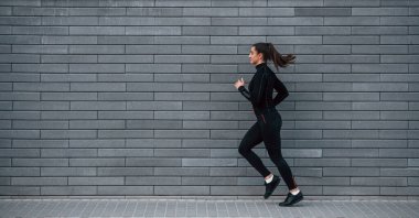 Siyah spor kıyafetli genç bir kız gri duvarın yanında koşuyor..