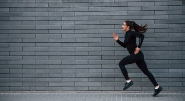 Young sportive girl in black sportswear running outdoors near gray wall.