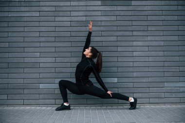 Siyah spor kıyafetli genç bir kız gri duvarın yanında yoga egzersizleri yapıyor..