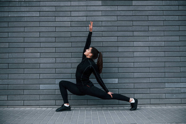 Young sportive girl in black sportswear outdoors doing yoga exercises near gray wall.