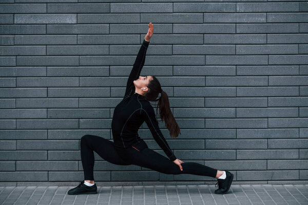 Young sportive girl in black sportswear outdoors doing yoga exercises near gray wall.