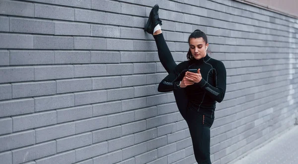 Young Sportive Girl Phone Black Sportswear Doing Legs Stretching Outdoors —  Fotos de Stock