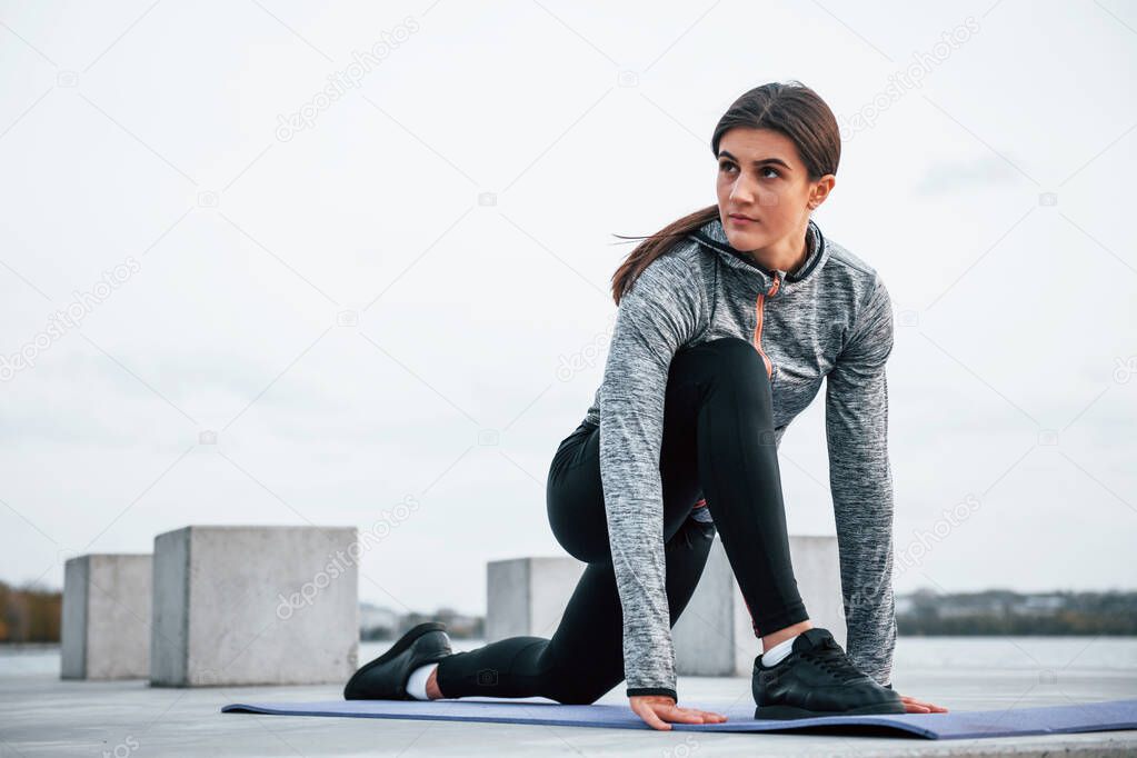 Young sportive girl doing yoga exercises on fitness mat outdoors near lake at daytime.