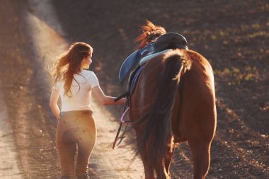 Koruyucu şapkalı genç bir kadın güneşli bir günde atıyla tarım alanında yürüyor..
