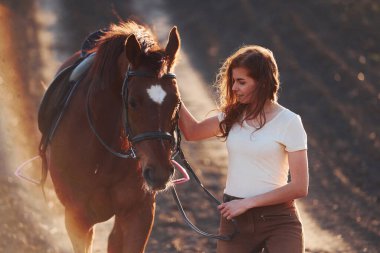 Koruyucu şapkalı genç bir kadın güneşli bir günde atıyla tarım alanında yürüyor..
