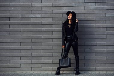 Beautiful brunette with curly hair and in black clothes holding bag outdoors near wall.