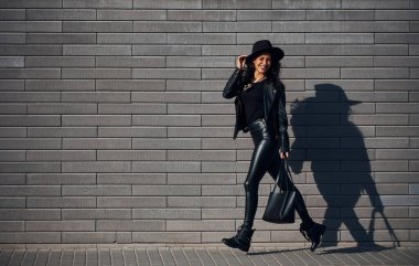 Kıvırcık saçlı, siyah elbiseli güzel bir esmer duvarın yanında yürür..