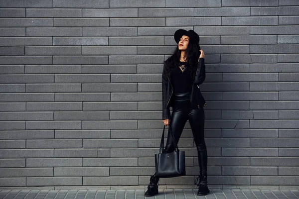 巻き毛と黒の服で美しいブルネットは壁の近くに屋外でバッグを保持 — ストック写真