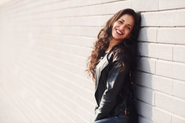 Potrait of cheerful brunette with curly hair and in black clothes leaning on the wall.