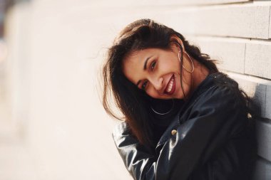 Potrait of cheerful brunette with curly hair and in black clothes leaning on the wall.