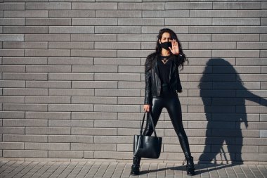 Durma hareketini elle gösteriyor. Koruyucu maskeli. Güzel esmer, kıvırcık saçlı ve dışarıda, duvarın yanında siyah elbiseli..