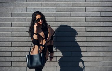 Beautiful brunette with curly hair and in black clothes talking by phone outdoors near wall.