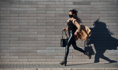 Koruyucu maskeli. Güzel esmer, kıvırcık saçlı, siyah elbiseli, duvarın yanında koşan..
