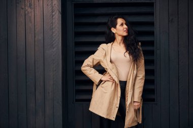 Woman with black curly hair standing against black wooden building exterior.