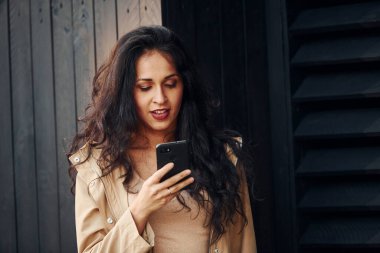 Siyah kıvırcık saçlı kadın siyah tahta binanın önünde duruyor ve telefon kullanıyor..