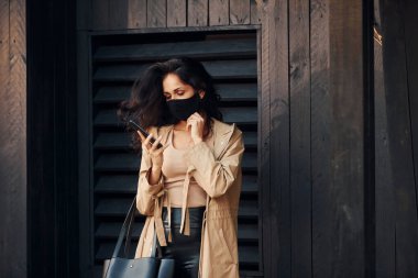 Koruyucu maskeli. Siyah kıvırcık saçlı kadın siyah tahta binanın önünde duruyor ve telefon kullanıyor..