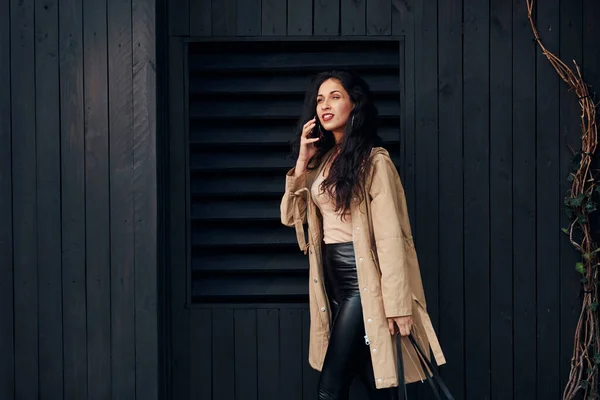 Woman with black curly hair standing against black wooden building exterior and using phone.