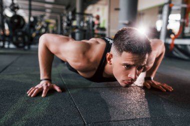 Doing push-ups. Young sportive strong man in black wear have workout day in gym.