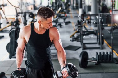 Dambılları yakmak. Siyah elbiseli genç sporcu güçlü adam spor salonunda antrenman yapıyor..