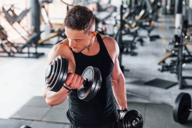 Dambılları yakmak. Siyah elbiseli genç sporcu güçlü adam spor salonunda antrenman yapıyor..