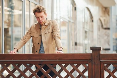 Elegant young man in formal classy clothes leaning on wooden fence outdoors in the city.