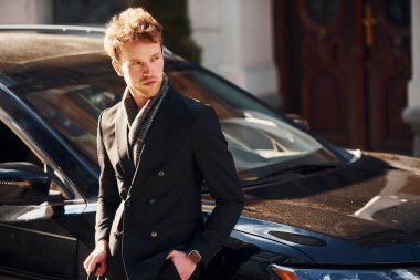 Standing near black car. Elegant young man in formal classy clothes outdoors in the city.