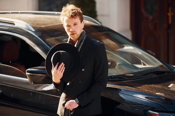 Standing near black car. Elegant young man in formal classy clothes outdoors in the city.