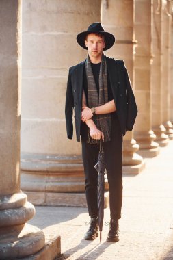 Elegant young man in black hat and clothes with umbrella in hands outdoors in the city.