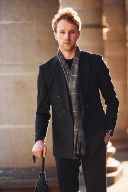 Holding umbrella. Elegant young man in formal classy clothes outdoors in the city.