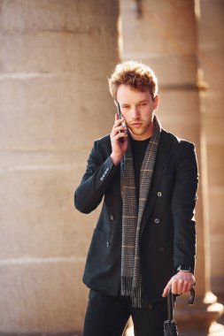 Şık giyinmiş zarif bir genç adam şehrin dışında telefonun başında dikiliyor ve konuşuyor..