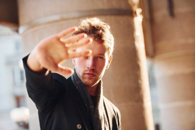Shows stop gesture by the hand. Elegant young man in formal classy clothes outdoors in the city.