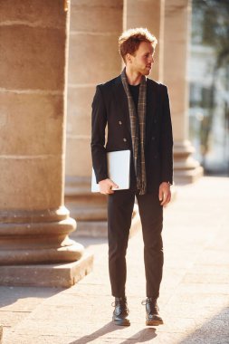 Ellerinde dizüstü bilgisayarla. Gözlüklü zarif genç adam şehrin dışında şık kıyafetler giyiyor..