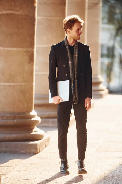 Ellerinde dizüstü bilgisayarla. Gözlüklü zarif genç adam şehrin dışında şık kıyafetler giyiyor..