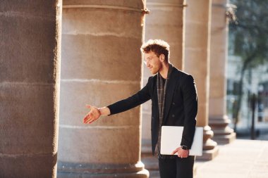 El hareketlerini gösteriyor. Ellerinde dizüstü bilgisayarla. Gözlüklü zarif genç adam şehrin dışında şık kıyafetler giyiyor..