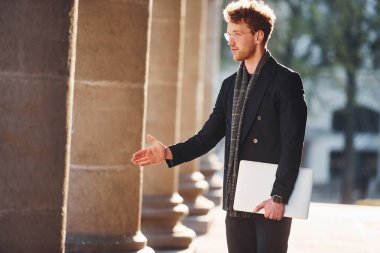 El hareketlerini gösteriyor. Ellerinde dizüstü bilgisayarla. Gözlüklü zarif genç adam şehrin dışında şık kıyafetler giyiyor..