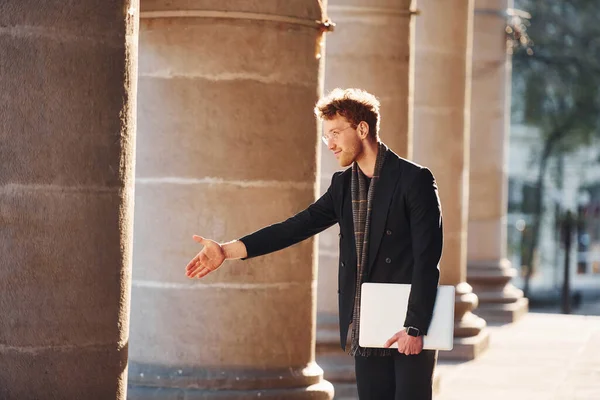 El hareketlerini gösteriyor. Ellerinde dizüstü bilgisayarla. Gözlüklü zarif genç adam şehrin dışında şık kıyafetler giyiyor..