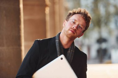 Güneşli bir hava. Ellerinde dizüstü bilgisayarla. Gözlüklü zarif genç adam şehrin dışında şık kıyafetler giyiyor..