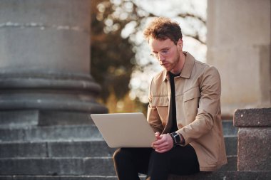 Dizüstü bilgisayarla merdivenlerde oturmak. Şehrin dışında şık kıyafetler içinde zarif bir genç adam..
