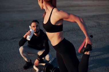 Adam oturur ve elinde bir şişe su tutar. Kadın ve erkeğin akşam vakti yolda spor günleri olur..