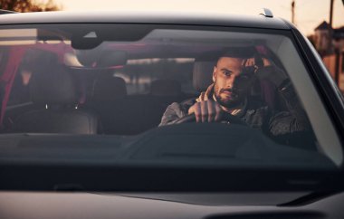 Moda giysileri içinde arabasıyla gezen yakışıklı, tıraşsız bir adamın ön görüntüsü..