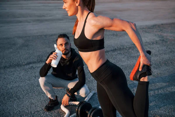 Adam oturur ve elinde bir şişe su tutar. Kadın ve erkeğin akşam vakti yolda spor günleri olur..