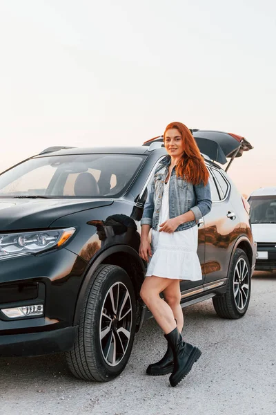 Redhead woman in skirt standing near black automobile on the road.
