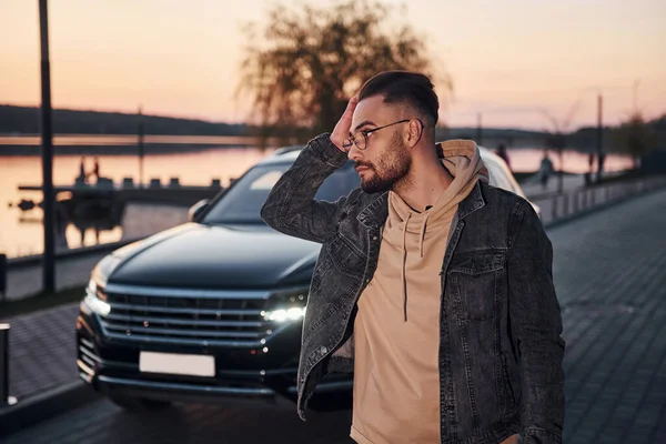 Şık giyinmiş yakışıklı, tıraşsız bir adam akşam vakti siyah arabasının yanında yürüyor..