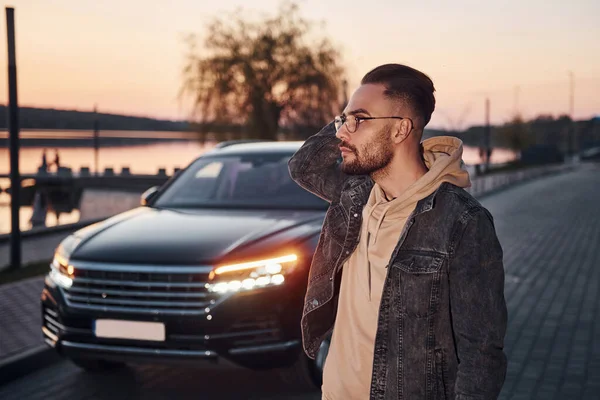 Şık giyinmiş yakışıklı, tıraşsız bir adam akşam vakti siyah arabasının yanında yürüyor..