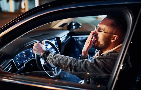 Şık giyinmiş, traşsız, yorgun ve yakışıklı bir adam otomobilini sürüyor..