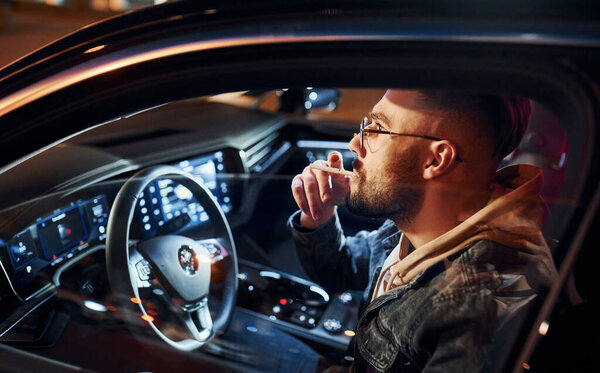 Курю сигарету. Красивый небритый мужчина в модной одежде на своем автомобиле.