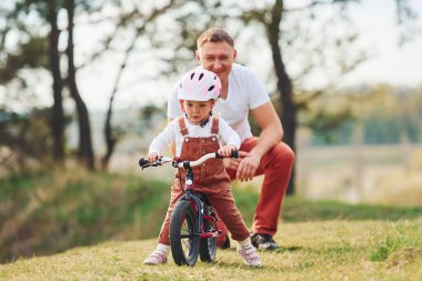 Father in white shirt teaching daughter how to ride bicycle outdoors. clipart