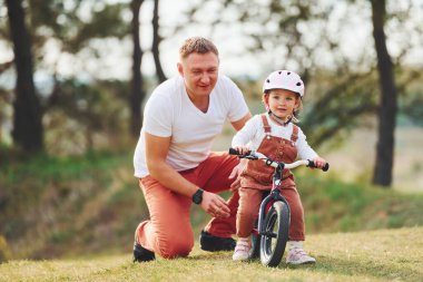 Beyaz gömlekli baba kızına dışarıda bisiklet sürmeyi öğretiyor..